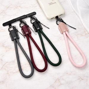 Puede incluir: Un conjunto de correas de muñeca para teléfono de colores gris, burdeos, verde y rosa. Cada correa presenta un lazo de cordón tejido unido a un adorno de cuero con un anillo metálico y un conector negro para el teléfono.