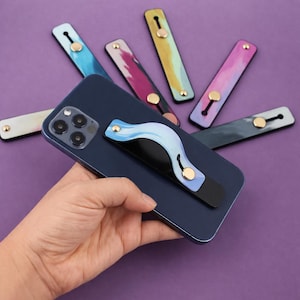 Op de afbeelding: Een marineblauwe smartphone met een blauwe en zwarte telefoonhouder bevestigd, vastgehouden door een hand. Verschillende kleurrijke telefoonhouders met aquarelontwerpen in tinten blauw, roze, geel en grijs zijn gerangschikt op een paarse achtergrond.