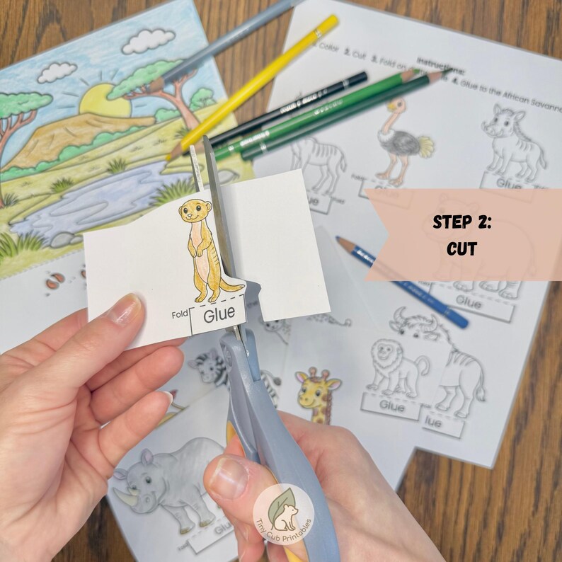 May include: A pair of grey scissors cutting a paper cutout of a meerkat with the text Glue and Fold. In the background, there are printed activity sheets featuring African savanna animals, colored pencils, and a text box labeled STEP 2: CUT.