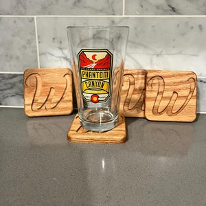 May include: A clear pint glass with a colorful Phantom Canyon Brewing Co. logo, resting on a wooden coaster. Three additional wooden coasters with a cursive "W" are visible. The coasters and glass are on a gray countertop.