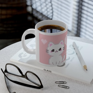 May include: White ceramic mug featuring a pixelated cat design on a pink backdrop. The mug is filled with dark coffee and is placed on a white surface with a pencil, glasses, and paperclips. The mug has a white handle.