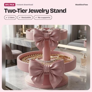 May include: A two-tier jewellery stand in a soft pink colour, featuring a central pillar and two circular trays. Each tier is decorated with a large, sculpted pink bow on the front. The stand holds various gold chains, bracelets, and rings.