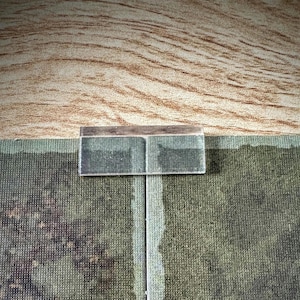 Op de afbeelding: Een klein, rechthoekig, helder glazen object met een donkere rand, rustend op een getextureerd oppervlak. Het object lijkt een klein, transparant paneel of een stuk glas te zijn. De achtergrond heeft een houtnerfpatroon en een groen, getextureerd oppervlak.