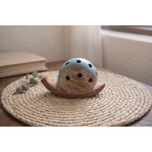 May include: A ceramic snail-shaped incense holder. The snail's shell is light blue with dark holes, and the body is a warm brown. The holder sits on a woven mat, suggesting a natural, earthy aesthetic. The design is simple and elegant.