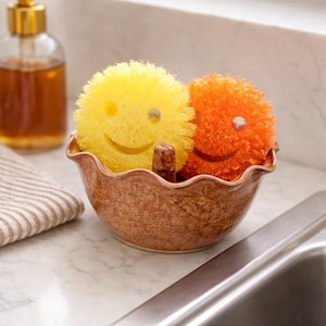 May include: A decorative brown ceramic bowl with a wavy rim holds two smiling face scrubbies. One scrubby is yellow, the other is orange. A brown ceramic divider separates the scrubbies. A glass soap dispenser is in the background.
