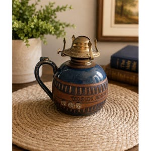 May include: A ceramic oil lamp with a rounded blue and brown body featuring a handle and a metallic brass burner assembly. The base is embossed with the text "GOD IS LIGHT, AND IN HIM IS NO DARKNESS" and decorative floral patterns around the center.