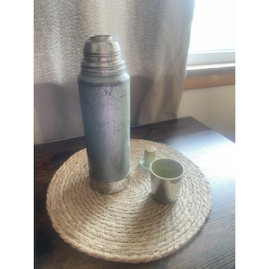 Può includere: Un thermos vintage color argento con tazza e tappo abbinati. Il thermos è cilindrico con coperchio e base argentati. La tazza e il tappo sono piccoli e argentati. Gli oggetti sono su un sottobicchiere rotondo e intrecciato.