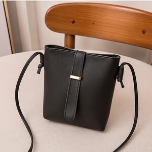 Puede incluir: Bolso bandolera negro con forma rectangular y una correa larga y fina. El bolso tiene un cierre de solapa con un acento metálico dorado. El bolso está hecho de un material liso y está sobre una silla.