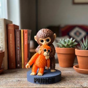 May include: A whimsical figurine featuring two monkey figures. One is brown and hugging an orange monkey. The base is blue and has the word "PUNCH" in black. The figurine is on a wooden surface.