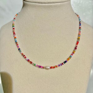 May include: A colorful beaded necklace with a central white pearl. The necklace features a variety of small, round beads in shades of red, orange, yellow, green, blue, and purple. The necklace is displayed on a mannequin.