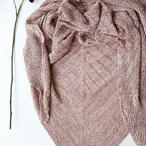 Puede incluir: Un chal tejido en tonos malva y beige, con un diseño geométrico. El chal está colocado sobre una superficie blanca, con una ramita de flores moradas oscuras en la esquina superior izquierda. El chal está hecho de hilo suave.