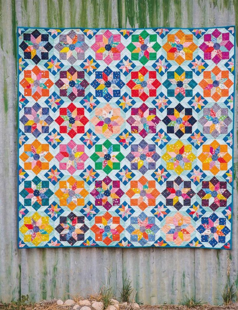 Puede incluir: Una colcha rectangular con un patr&oacute;n repetitivo de bloques de estrellas multicolores en tonos rojo, naranja, amarillo, verde, azul, morado y rosa. Las estrellas est&aacute;n sobre un fondo azul claro con un borde de color azul s&oacute;lido.