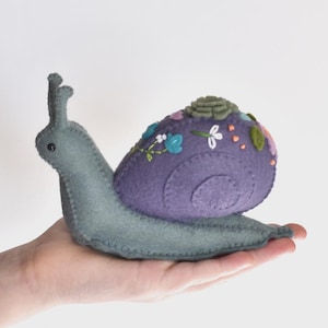 May include: A felt snail figurine with a light gray body and a dark purple shell. The shell is decorated with embroidered floral patterns in shades of blue, white, and green, featuring a small green succulent-like accent on the top.