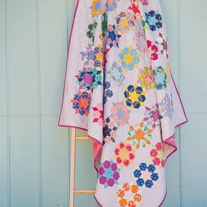 Puede incluir: Una colcha con fondo lavanda claro y un patr&oacute;n repetitivo de motivos florales y geom&eacute;tricos en forma de estrella de varios colores. La colcha tiene un ribete morado vibrante en los bordes y est&aacute; colgada sobre una escalera de madera.