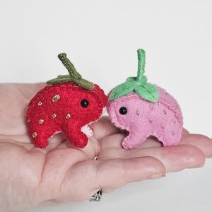 May include: Two small felted plush figures shaped like frogs with strawberry features. One frog is bright red with a dark green stem and gold embroidered seeds, while the other is light pink with a bright green stem and light pink embroidered seeds.