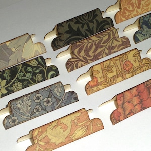 May include: A collection of decorative paper tabs with floral and botanical patterns in various colors, including shades of brown, green, and orange. Each tab has a small, white, rectangular extension for insertion. The tabs are arranged diagonally across a white surface.