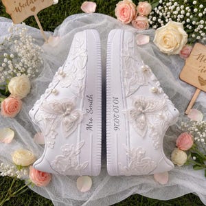 May include: A pair of white sneakers decorated with lace butterfly appliques, pearls, and floral embroidery. The left shoe features the text 'Mrs. Smith' and the right shoe features the date '10.10.2026' printed on the side of the rubber soles.