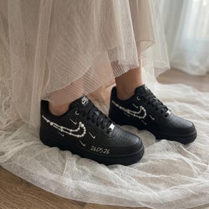 Könnte beinhalten: Schwarze Nike Air Force Sneaker mit weißen Perlenverzierungen entlang des seitlichen Swoosh-Logos, zusätzlichen kleinen gestickten Swoosh-Symbolen und dem numerischen Text 260526, der in Weiß auf dem vorderen Bereich des Schuhs aufgedruckt ist.
