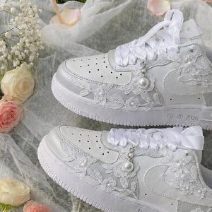 May include: A pair of white trainers featuring intricate white floral lace overlays, satin ribbon laces, and small pearl accents. The side of one shoe displays the text 05 20 2026, while the other shoe features the text Mrs. Smith in cursive script.