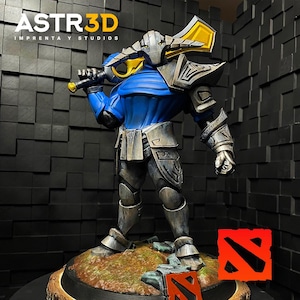 Puede incluir: Figura detallada de un personaje de Dota 2, con armadura azul y plateada y una gran arma amarilla y plateada. La base incluye el logotipo de Dota 2 y el texto "ASTR3D IMPRENTA Y STUDIOS".