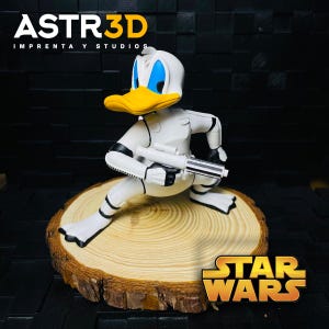 May include: A figurine of Donald Duck in a white Stormtrooper suit, holding a blaster. The duck has a yellow beak, blue eyes, and is posed on a wooden slice. The Star Wars logo is visible.