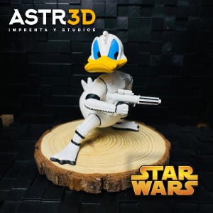 May include: A white and yellow Donald Duck figurine in a Stormtrooper outfit, holding a blaster. The figurine is on a wooden slice, with the Star Wars logo in the bottom right corner. The text "ASTR3D IMPRENTA Y STUDIOS" is at the top.