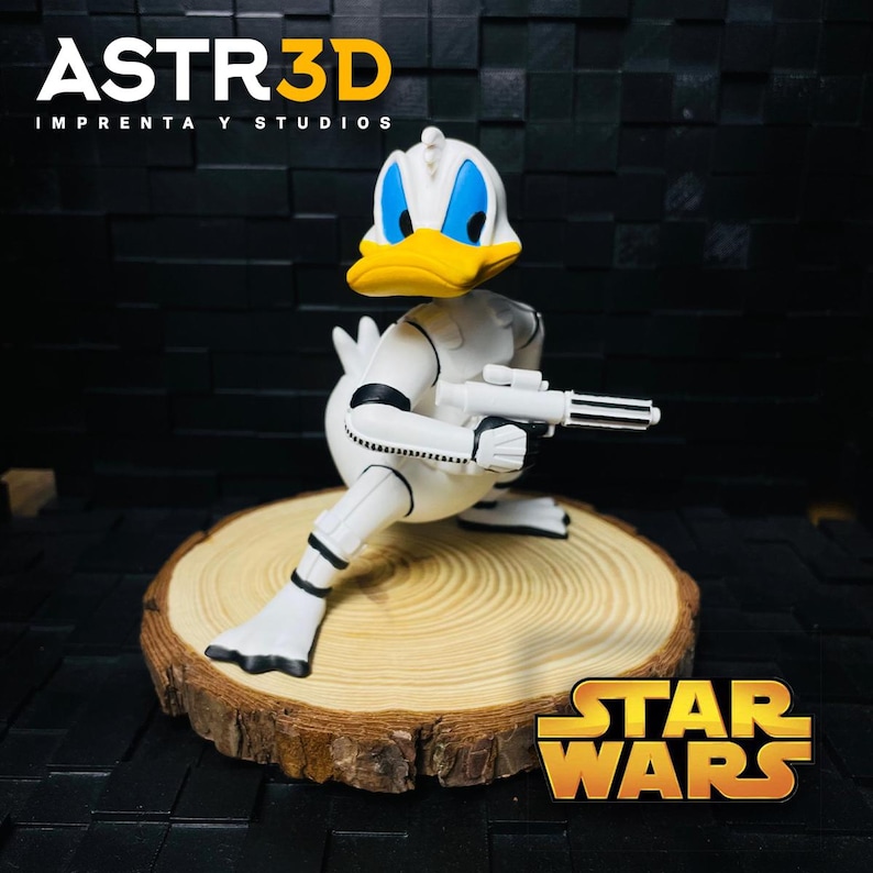 May include: A white and yellow Donald Duck figurine in a Stormtrooper outfit, holding a blaster. The figurine is on a wooden slice, with the Star Wars logo in the bottom right corner. The text "ASTR3D IMPRENTA Y STUDIOS" is at the top.