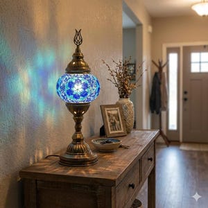 Puede incluir: Lámpara de mesa decorativa con pantalla de mosaico de vidrio en tonos azules y blancos. La lámpara tiene una base de metal color bronce y un remate en forma de tulipán. La lámpara está colocada sobre una mesa de madera en un pasillo.
