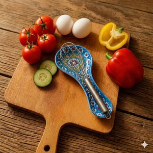 Puede incluir: Una tabla de cortar de madera con una variedad de ingredientes frescos. Incluye tomates rojos, rodajas de pepino, dos huevos blancos, un pimiento rojo y medio pimiento amarillo. Un reposacucharas azul y blanco con dibujos sostiene un batidor.