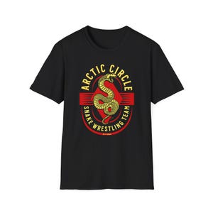 May include: Black t-shirt featuring a circular graphic with the text "Arctic Circle Snake Wrestling Team." The design includes a yellow snake and red accents. The shirt is made of a soft material.