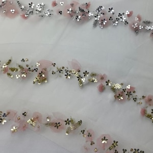 May include: Close-up of three decorative floral trims. Each trim features a delicate arrangement of pink fabric flowers, silver and gold sequins, and small, dark-colored accents. The trims are set against a white, sheer background.