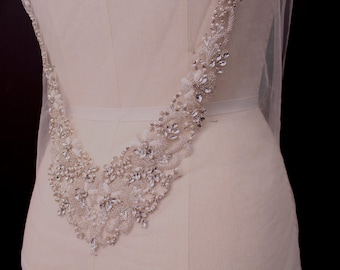 May include: A bridal bodice detail featuring a V-shaped design. The embellishment is made of white beads and clear crystals, creating a sparkling effect. The design is attached to a sheer fabric.
