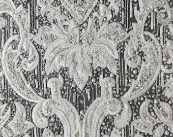 May include: Close-up of white embroidered fabric with a floral pattern. The design features intricate details with beads and sequins, creating a textured and ornate appearance. The fabric has a dark background, enhancing the brightness of the white embroidery.