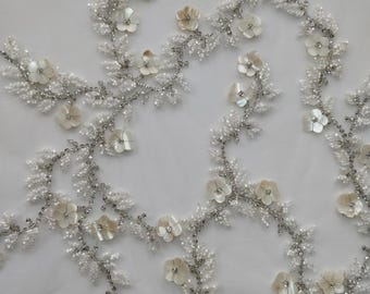 May include: Close-up of white floral lace fabric. The lace features delicate, off-white flowers with iridescent centers, interspersed with silver beaded vines and tiny white beads. The design is on a sheer white background.