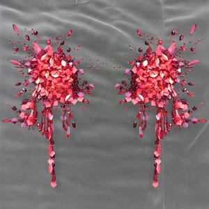 Może przedstawiać: Dwie symetryczne, bogato zdobione aplikacje z czerwonych cekinów i koralików. Każda aplikacja ma centralną grupę czerwonych cekinów, z kaskadowymi pasmami koralików i rozproszonymi akcentami. Aplikacje są umieszczone na szarym tle z siatki.