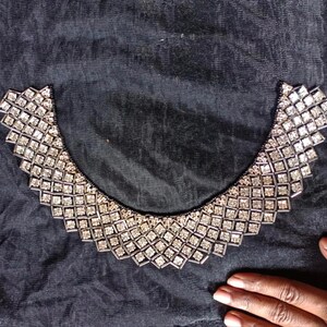 Op de afbeelding: Een decoratieve kraag met zilveren en heldere vierkante strass-steentjes. De kraag heeft een geometrisch patroon en is geplaatst op een donkere stof. De strass-steentjes glinsteren en vangen het licht.