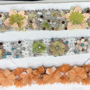 Op de afbeelding: Drie decoratieve bloemenborduursels. De bovenste rand heeft perzik- en groene stoffen bloemen met zilveren en iriserende kralen. De middelste rand heeft witte en groene bloemen met blauwe en heldere kralen. De onderste rand heeft oranje stoffen bloemen.