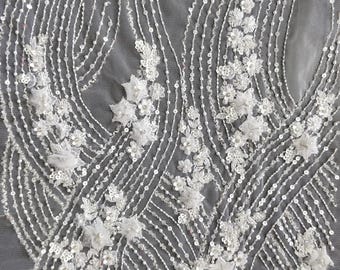 May include: Close-up of white embroidered fabric with a delicate floral and sequin design. The fabric features vertical and curved lines of sequins and small white flowers, creating a textured, elegant pattern. Ideal for bridal or formal wear.
