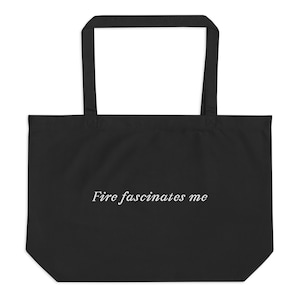 Könnte beinhalten: Schwarze Tragetasche mit langen Henkeln. Auf der Tasche steht in einer weißen, eleganten Schrift der Text "Fire fascinates me". Die Tasche ist aus einem robusten Material gefertigt und zum Tragen von Gegenständen bestimmt.