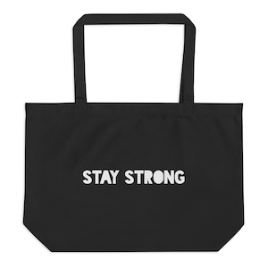 May include: Black tote bag with white text that reads "STAY STRONG". The bag has a rectangular shape with a large open top and two long handles. The bag is made of a sturdy material.
