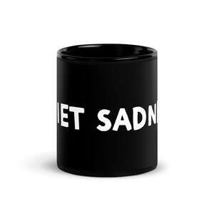May include: A black ceramic mug with the white text "QUIET SADNESS" printed on the side. The mug is cylindrical and appears to be empty. The mug is a solid black color.