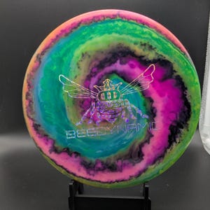 May include: A disc golf disc with a vibrant, swirling design in shades of green, pink, orange, and blue. It has a bee graphic and the text "BEEDYNAMIC". The disc is presented on a black stand.
