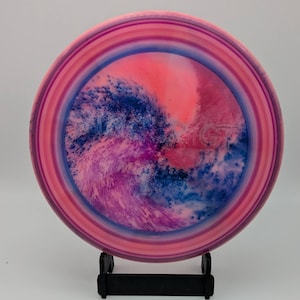 Op de afbeelding: Een ronde discgolfschijf met een levendig tie-dye patroon met wervelende tinten roze, magenta en diepblauw. De buitenrand vertoont concentrische ringen van licht- en donkerroze, terwijl in het midden de letters GT in reliëf zijn aangebracht.