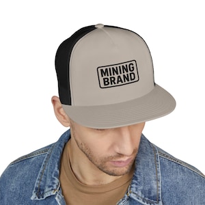Puede incluir: Gorra de camionero beige y negra con el texto "MINING BRAND" en un recuadro negro. La gorra tiene una visera curva y una parte trasera de malla.