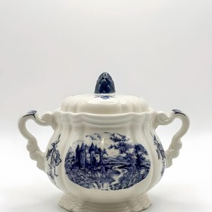 Peut inclure: Sucrier en céramique blanche avec un motif paysager bleu et blanc représentant un château, des arbres et une rivière. Le bol a deux anses et un couvercle assorti avec un bouton bleu foncé. Le bol mesure environ 15 cm de haut.