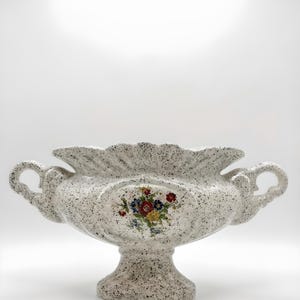 Peut inclure: Un vase en céramique blanc moucheté avec un socle et deux poignées. Le vase présente un motif floral au centre et un bord festonné. Le vase mesure environ 30 cm de large.