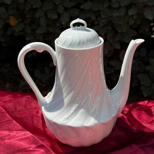Peut inclure: Une cafetière en porcelaine blanche avec un couvercle et une poignée incurvée. Le pot a un motif tourbillonnant texturé et un long bec. La cafetière est posée sur une surface rouge.