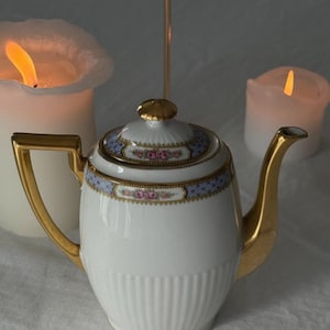 Puede incluir: Una tetera de porcelana blanca con detalles dorados, incluyendo el asa, el pico y la tapa. La tapa y el borde presentan un diseño floral azul y rosa. El fondo es suavemente borroso, con velas encendidas.