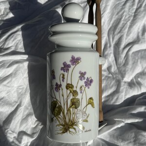 Peut inclure: Pot en céramique blanc avec couvercle et bouton sphérique, orné d'un motif floral de violettes violettes et de feuilles vertes. Deux cuillères en bois sont derrière le pot. Le pot porte l'inscription "Viola odorata".