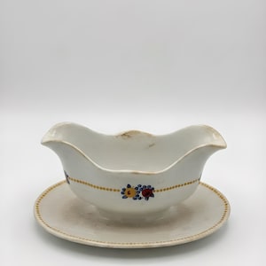 Puede incluir: Una salsera de cerámica blanca con su platillo a juego. La salsera tiene un borde festoneado y está decorada con una línea punteada dorada y detalles florales en amarillo, azul y rojo. Un plato para servir vintage.